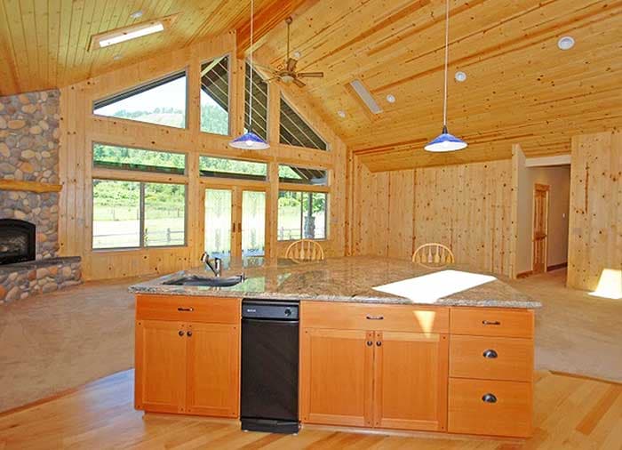 Interior view of a great room with stone fireplace, kitchen island with sink, and expansive windows with vaulted wood ceiling.