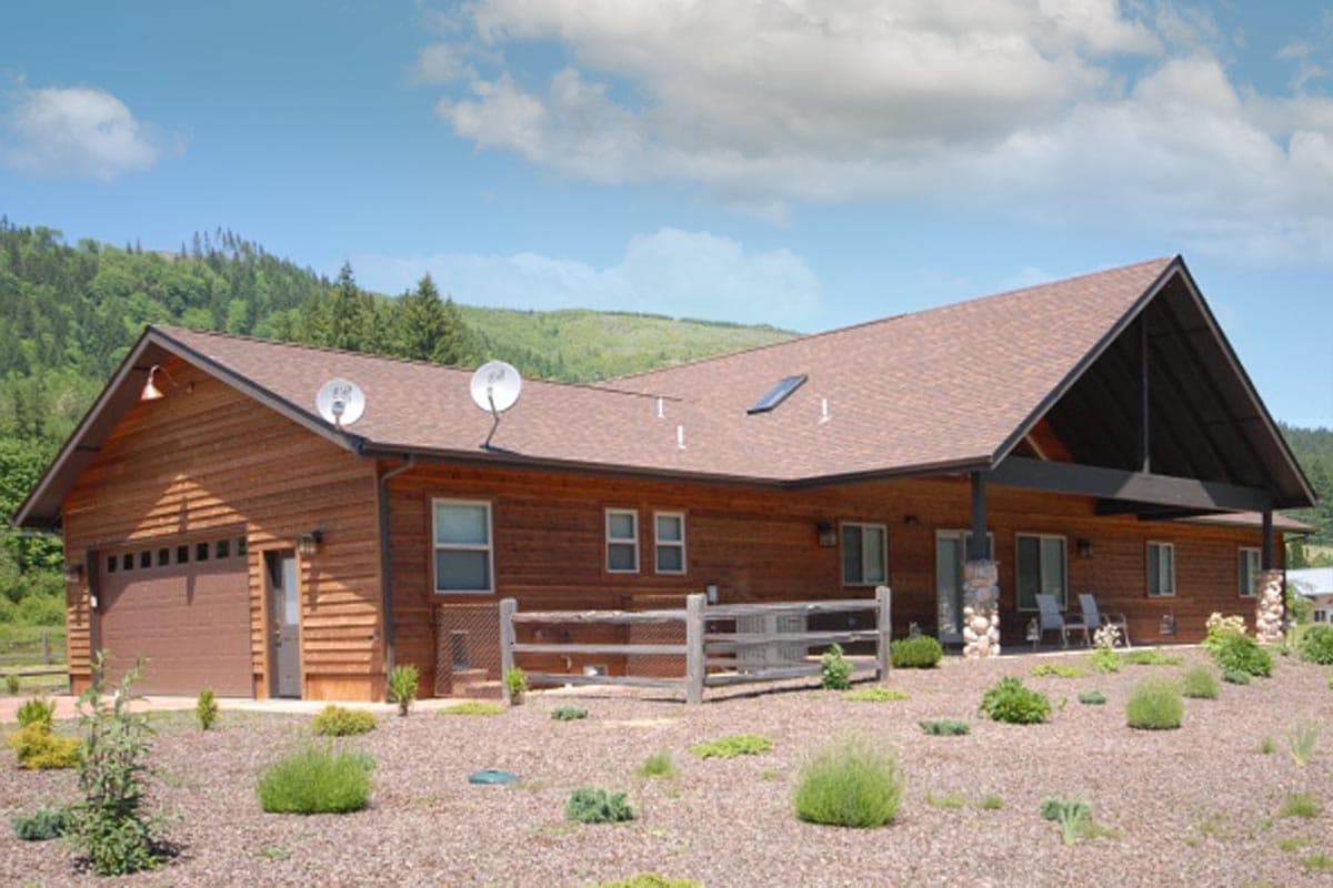 House plan exterior: Wood-sided ranch with attached garage, prominent gabled roof over covered patio, and satellite dishes.