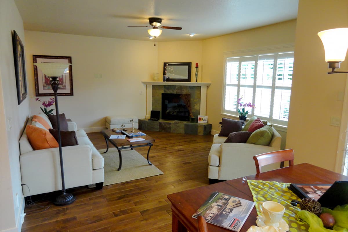 Living room with fireplace, wood floors, ceiling fan, and large window with shutters.