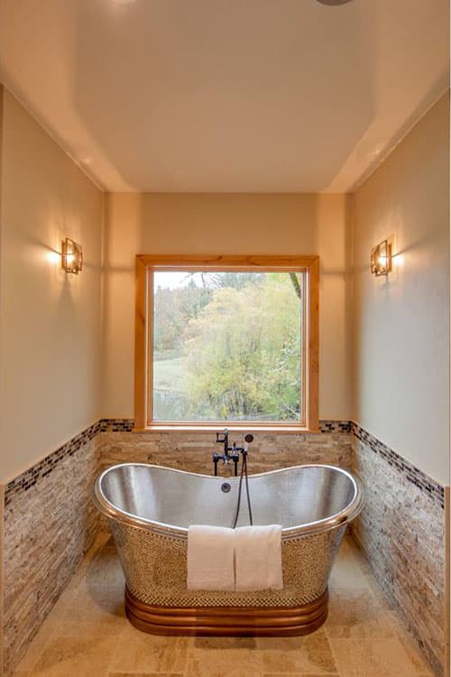 Bathroom with freestanding metal tub, stone tile wainscoting, and large window overlooking trees.