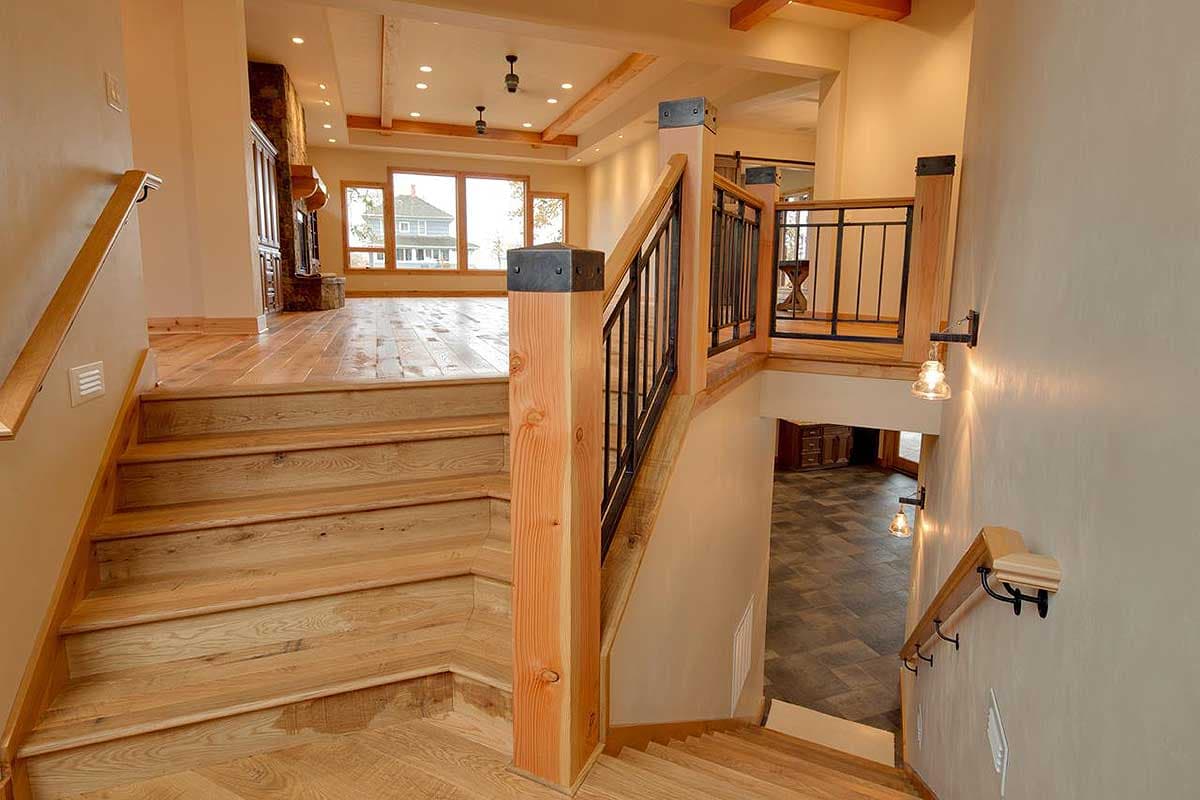 Interior view of a house showing a wooden staircase, exposed beams, stone fireplace, and large windows overlooking a home exterior.