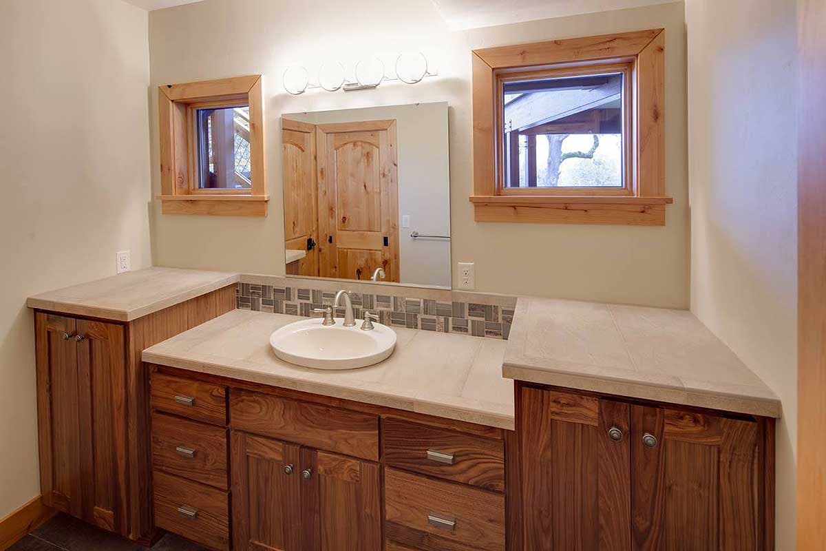 Bathroom vanity with a double sink, wood cabinetry, tile backsplash, and two small windows. Floor plan view.
