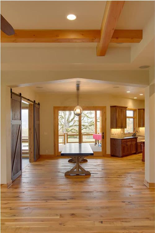 Interior view of a dining area with wood beam ceiling, barn doors, and view to kitchen and deck.