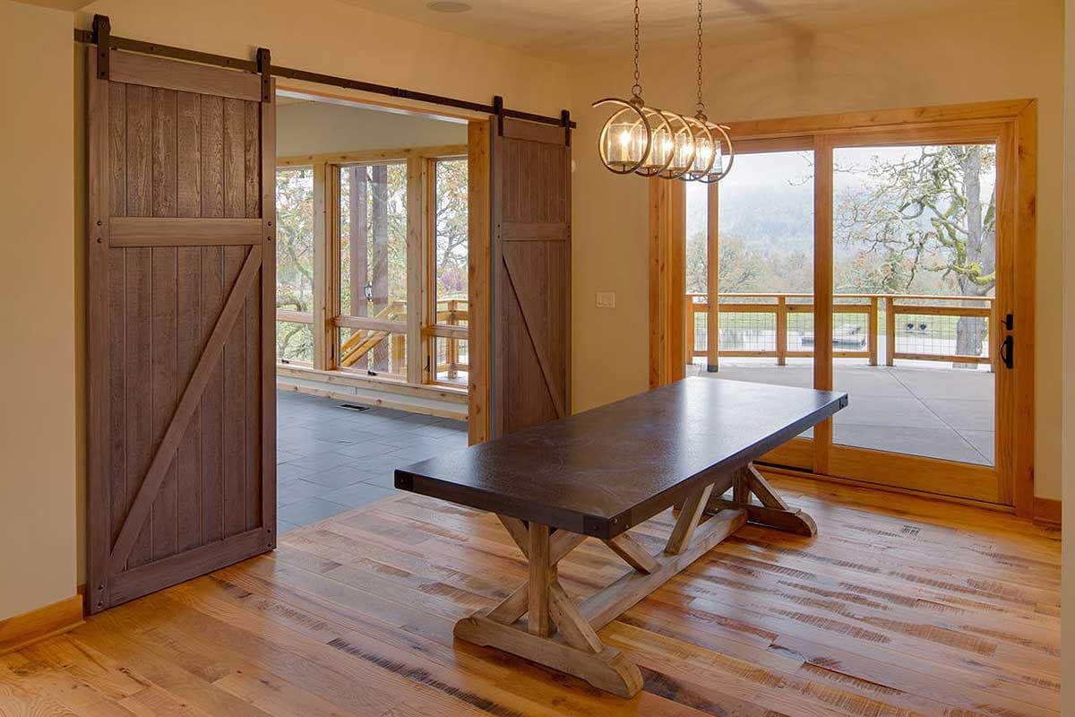 Dining area with wooden sliding barn doors, large windows, and deck access. Three globe pendant lights hang above a trestle table.