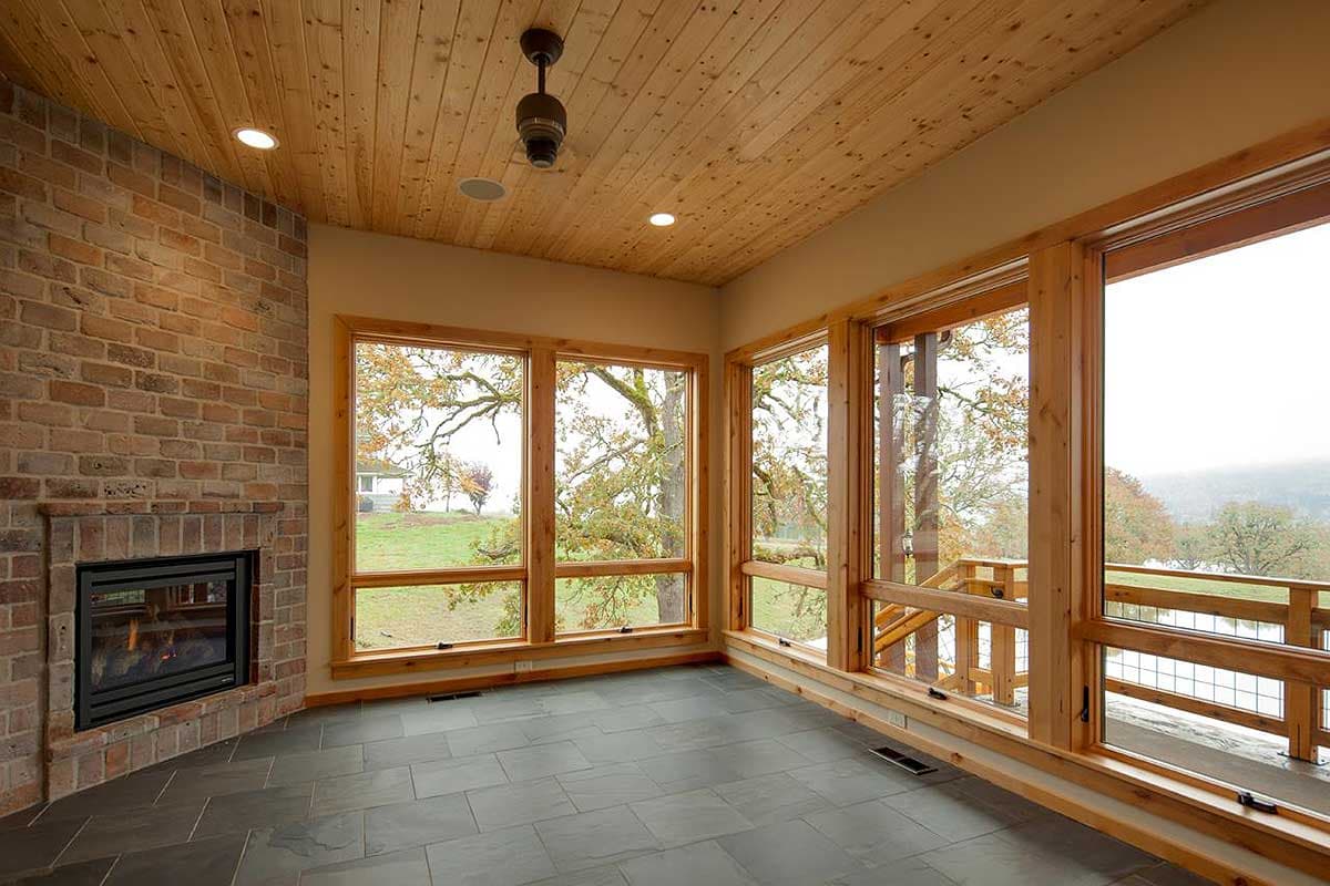 Interior view of a room with a brick fireplace, wood-paneled ceiling, large wood-framed windows, and slate tile flooring.