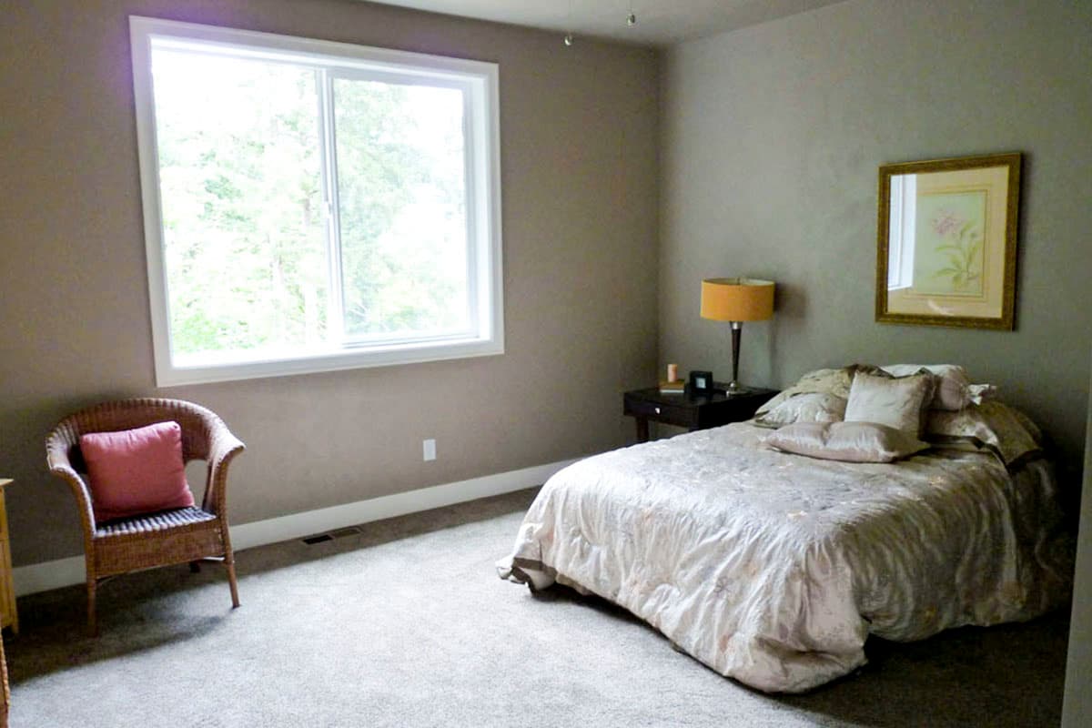 Bedroom with queen bed, bedside table, lamp, wicker chair with pink cushion, and large window overlooking trees.