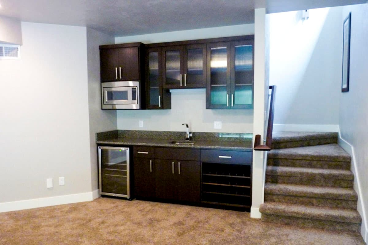 Wet bar with dark cabinetry, microwave, sink, wine rack, and built-in cooler adjacent to carpeted stairs.