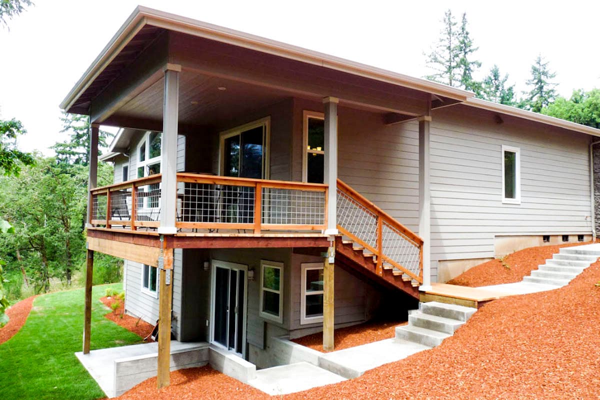 House plan exterior: Two-story modern home with a covered upper deck, wooden railings, and an external staircase leading to a lower level entrance.