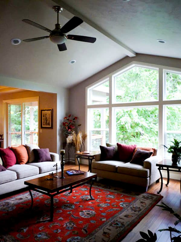 Living room with vaulted ceiling, ceiling fan, and large gable windows overlooking trees. Open to dining.