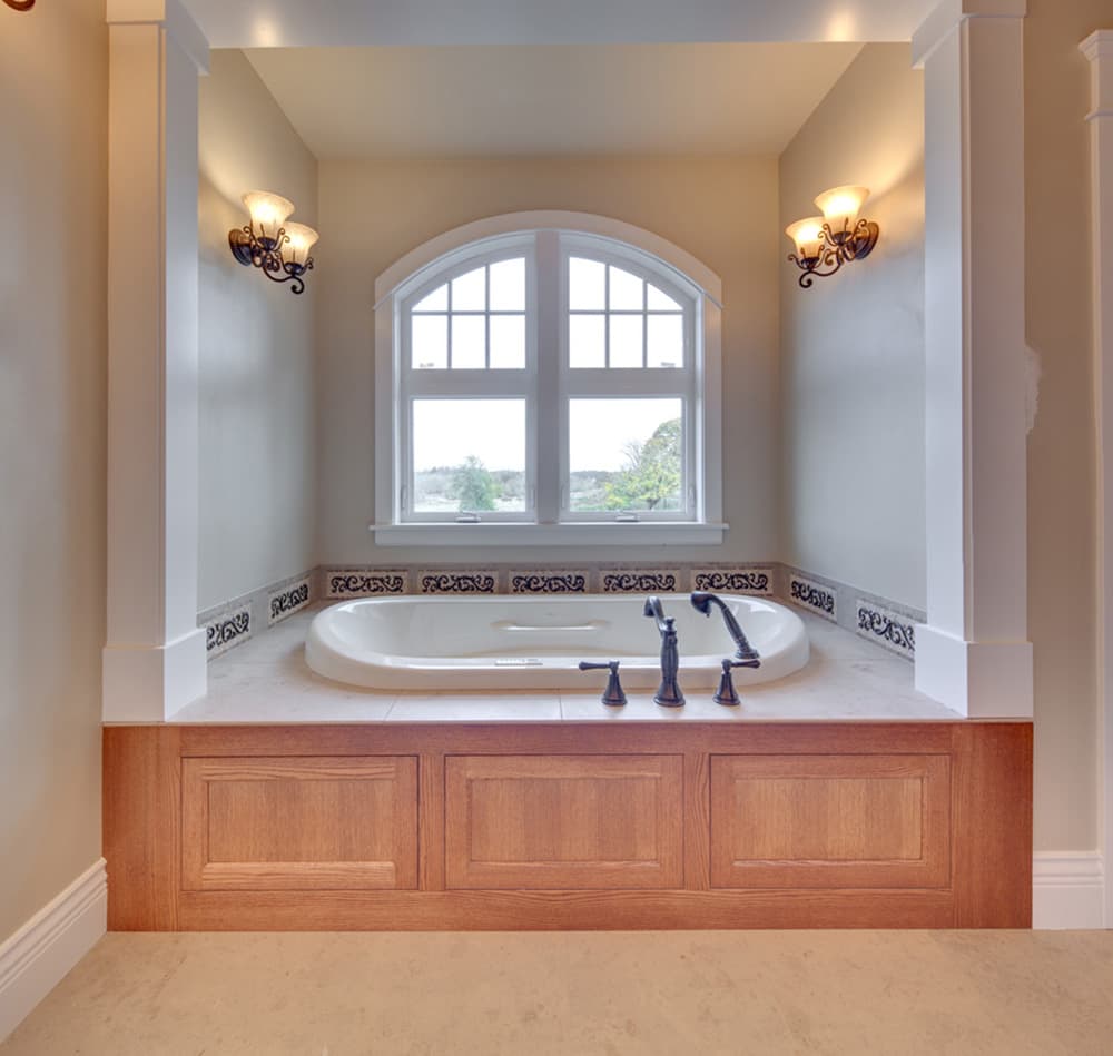 Bathroom with sunken soaking tub, arched window, ornate tile backsplash, and wood paneled base.