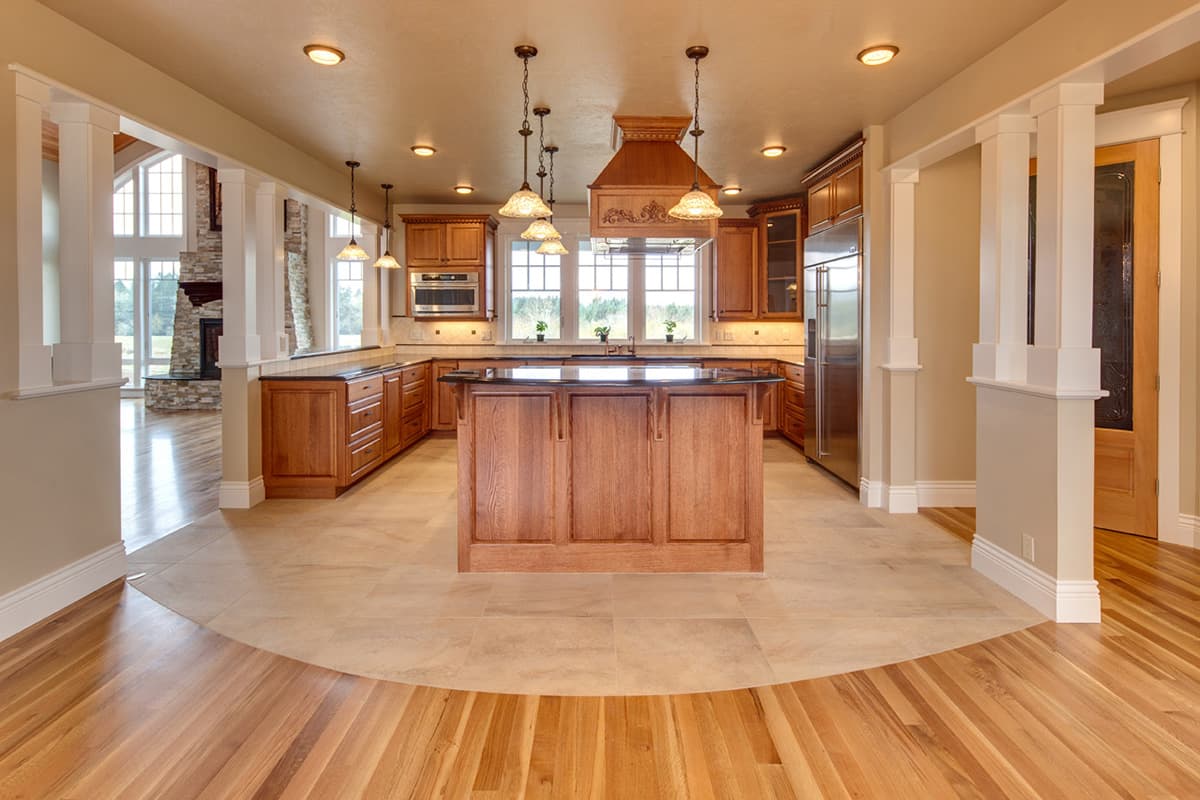 Kitchen interior with wooden cabinets, island, granite countertops, stainless steel appliances, and view into great room with stone fireplace.