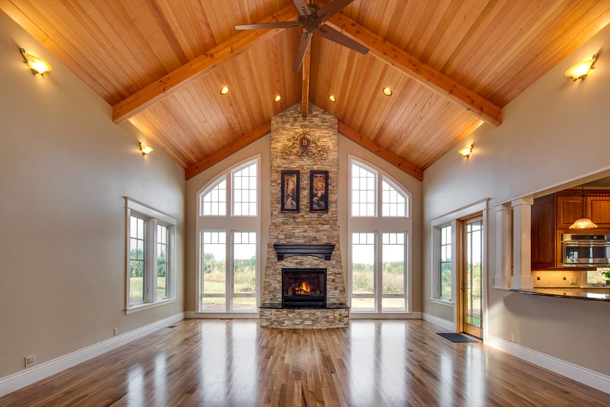Great room with vaulted wood ceiling, stone fireplace, and large arched windows overlooking landscape.