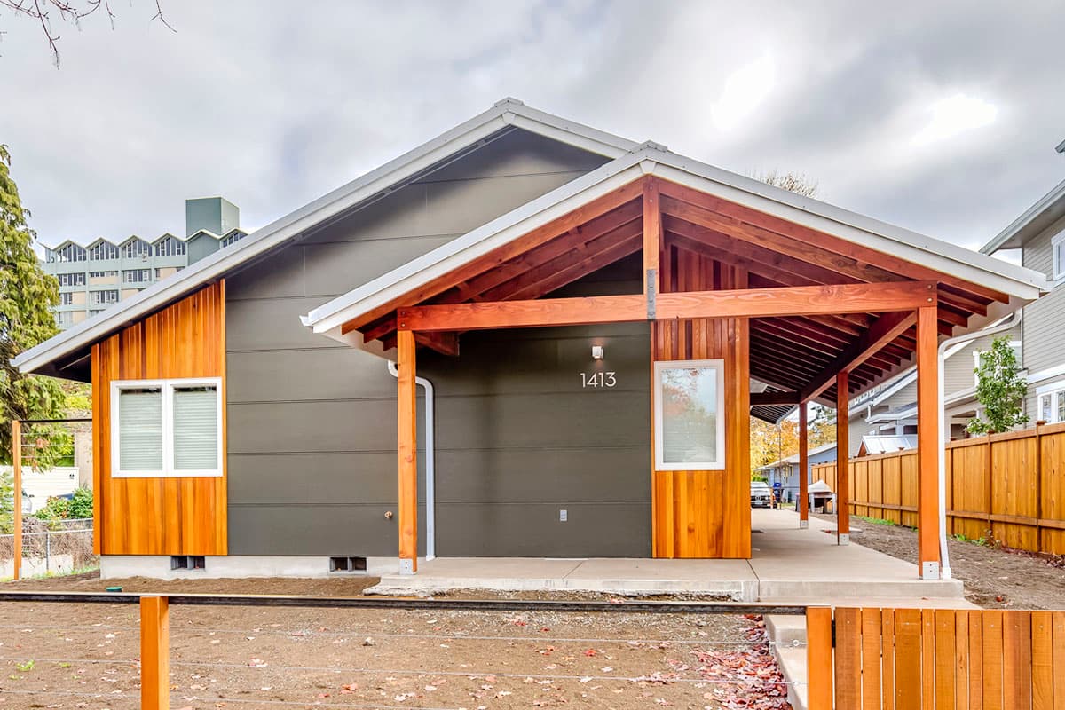 House plan exterior: one-story modern home with wood accents, prominent rafters on an open carport, and gable rooflines.
