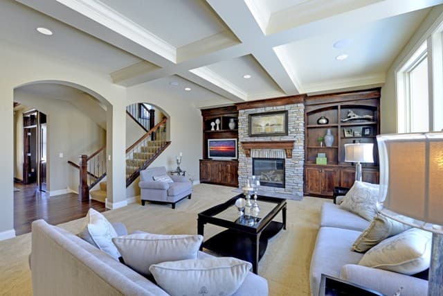 Living room with coffered ceiling, stone fireplace, built-in bookshelves, and view of arched staircase.