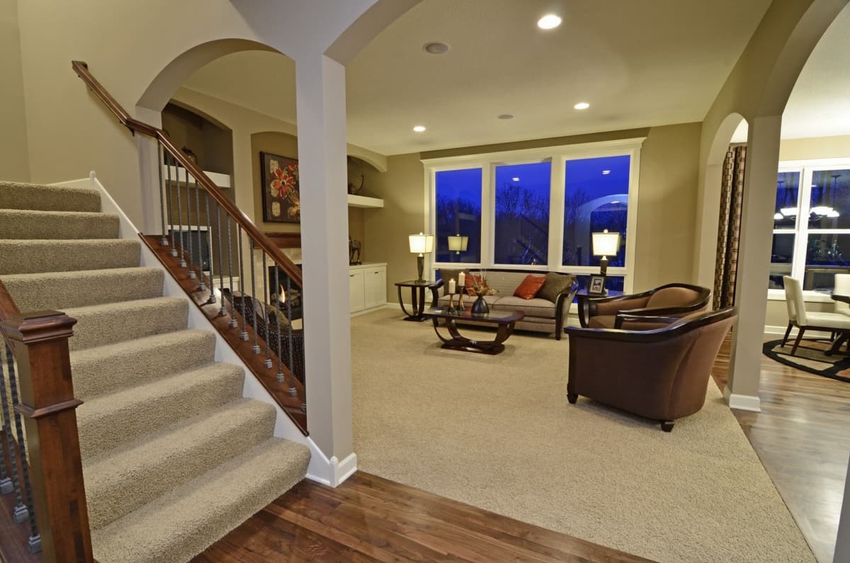 Interior view of a house plan with carpeted stairs, living room with large windows, and a dining area visible.