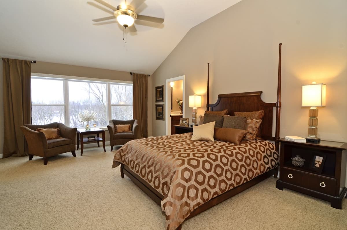 Primary bedroom with vaulted ceiling, four-poster bed, sitting area, and large windows overlooking winter trees.