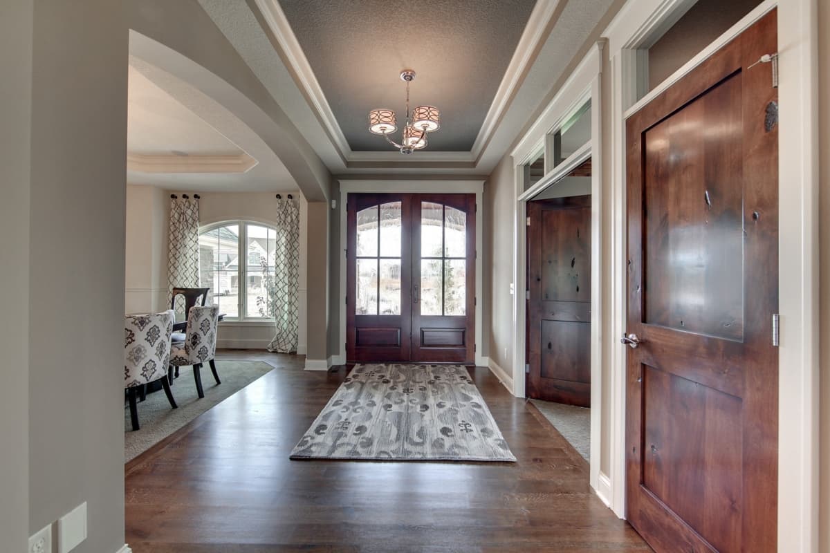 Interior view of a house entrance with double wooden doors, coffered ceiling, and archway leading to a dining area with a large window.