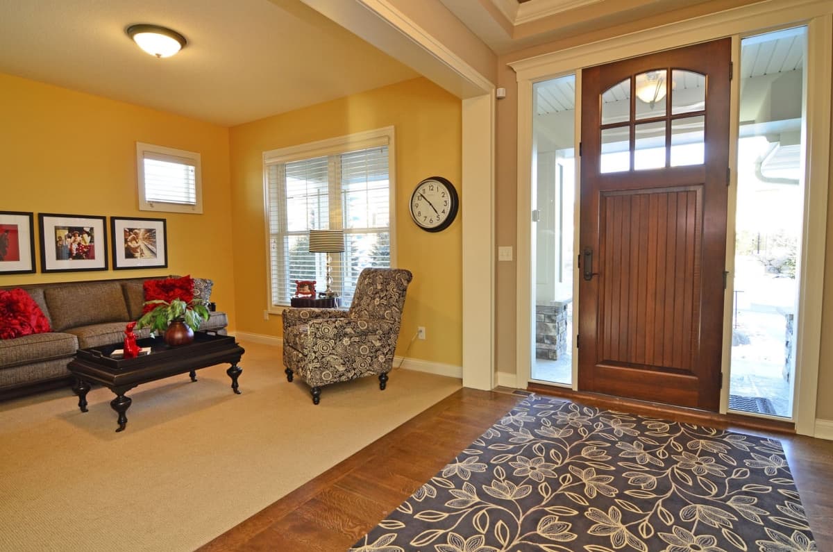 Interior view of a living room with yellow walls, sofa, patterned armchair, coffee table, and a wooden front door with glass sidelights.