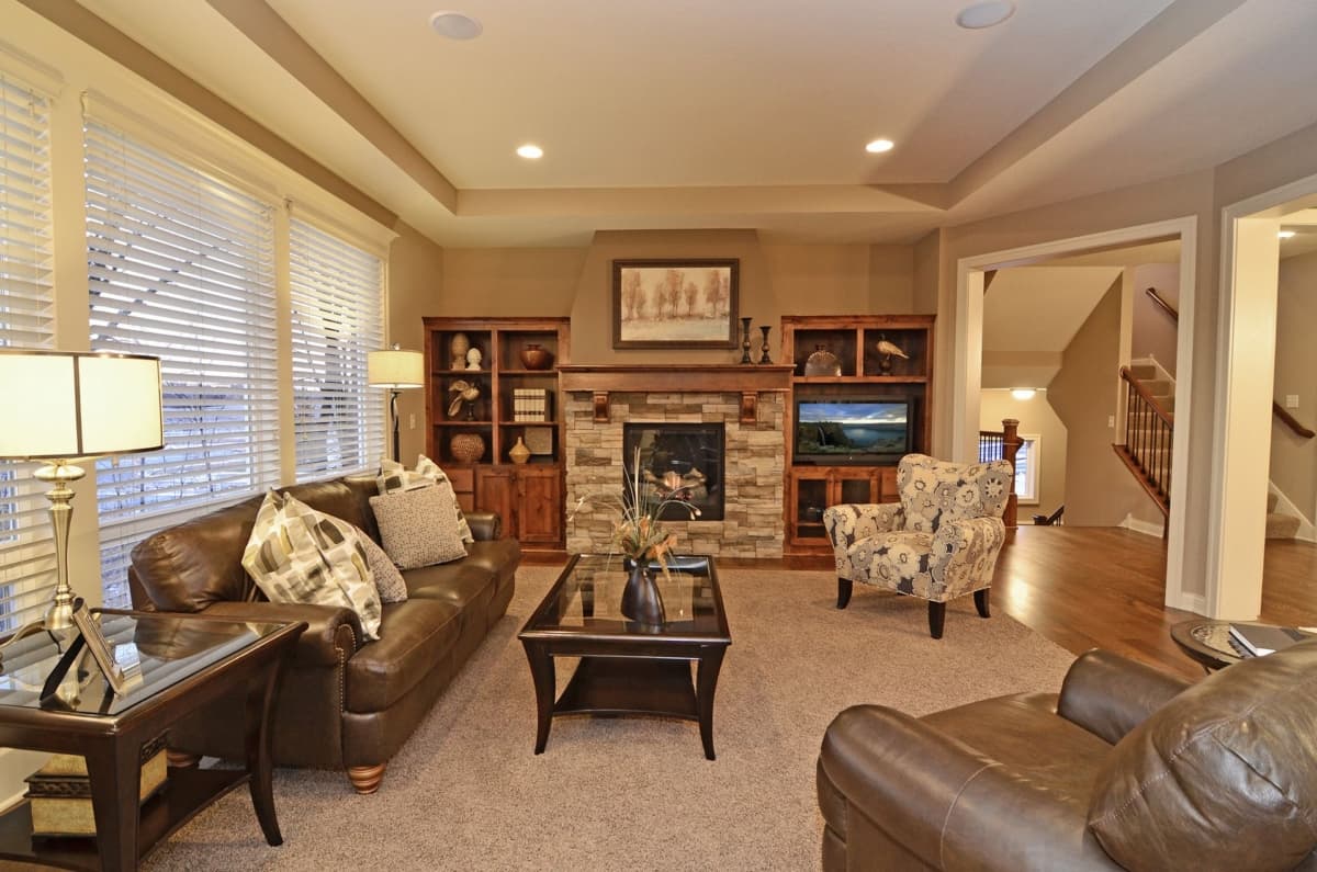 Interior view of a living room with a stone fireplace, built-in bookshelves, leather sofa, and open doorway to a staircase.