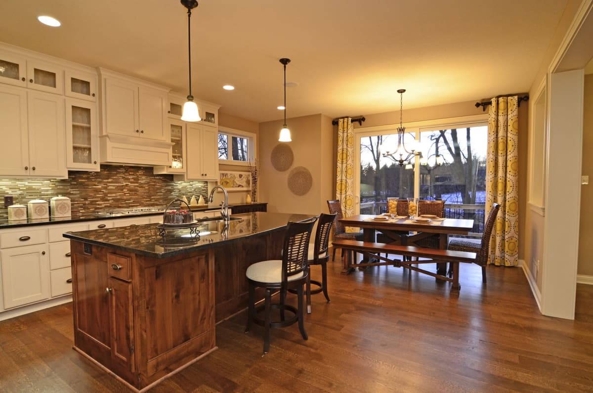 Kitchen with island and dining area. Features white cabinetry, granite countertops, mosaic tile backsplash, wood flooring, and large windows.