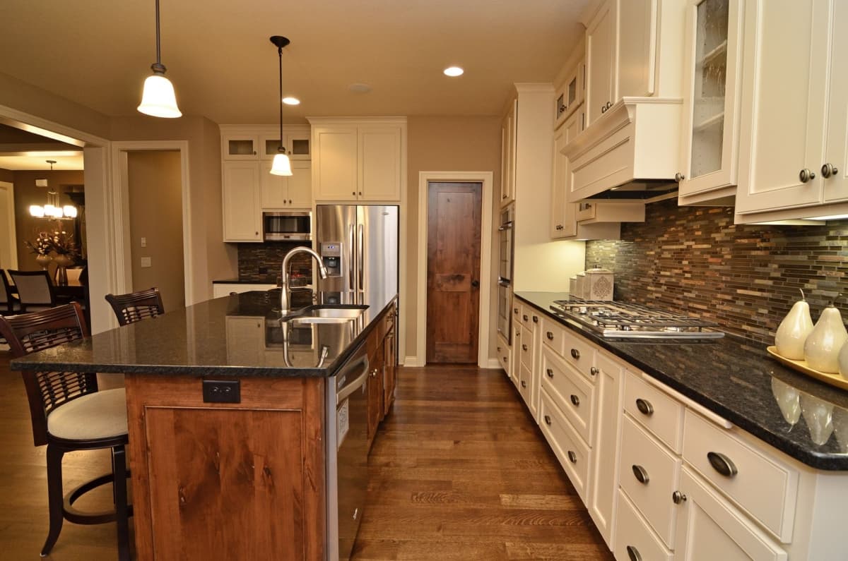 Kitchen interior with large island, granite countertops, white cabinets, stainless steel appliances, and a mosaic tile backsplash.