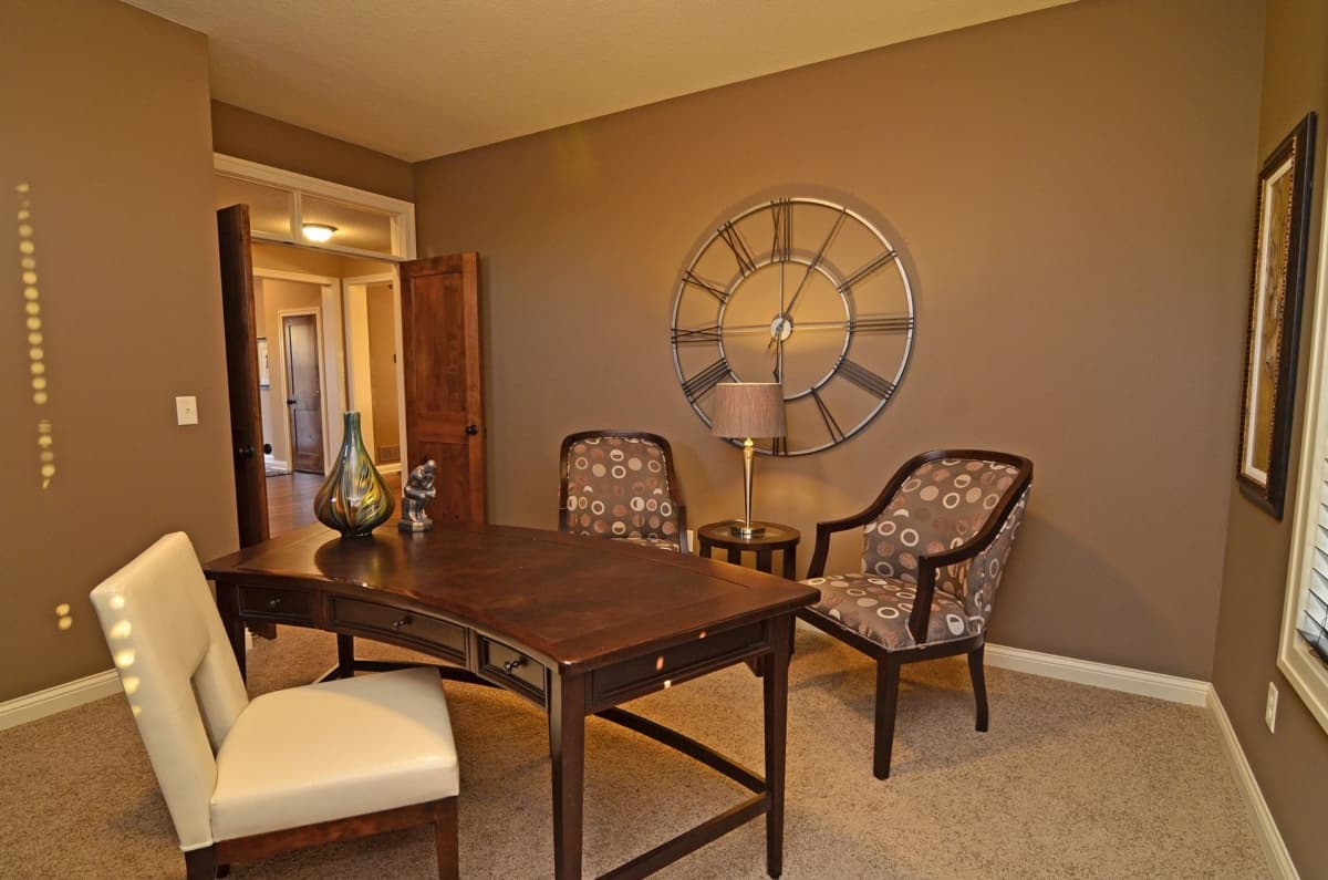 Office with wood desk, two patterned chairs, a large wall clock, and an open doorway revealing a hallway.