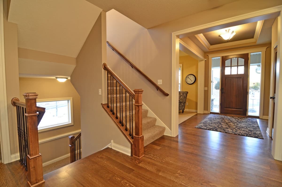 Interior view of a house entrance and staircase with wood flooring, wooden banister, carpeted stairs, and a coffered ceiling above the entryway.