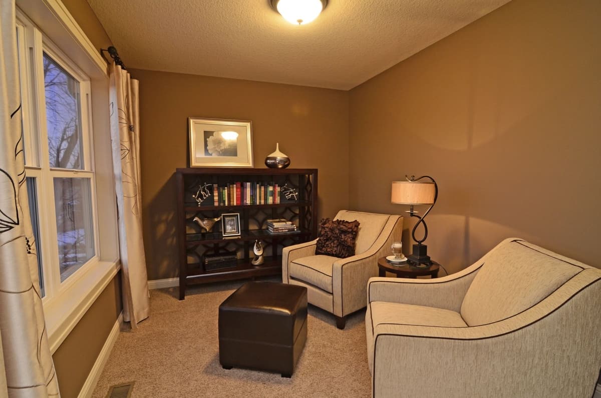 Interior view of a reading nook with two armchairs, an ottoman, a bookshelf, and a large window.