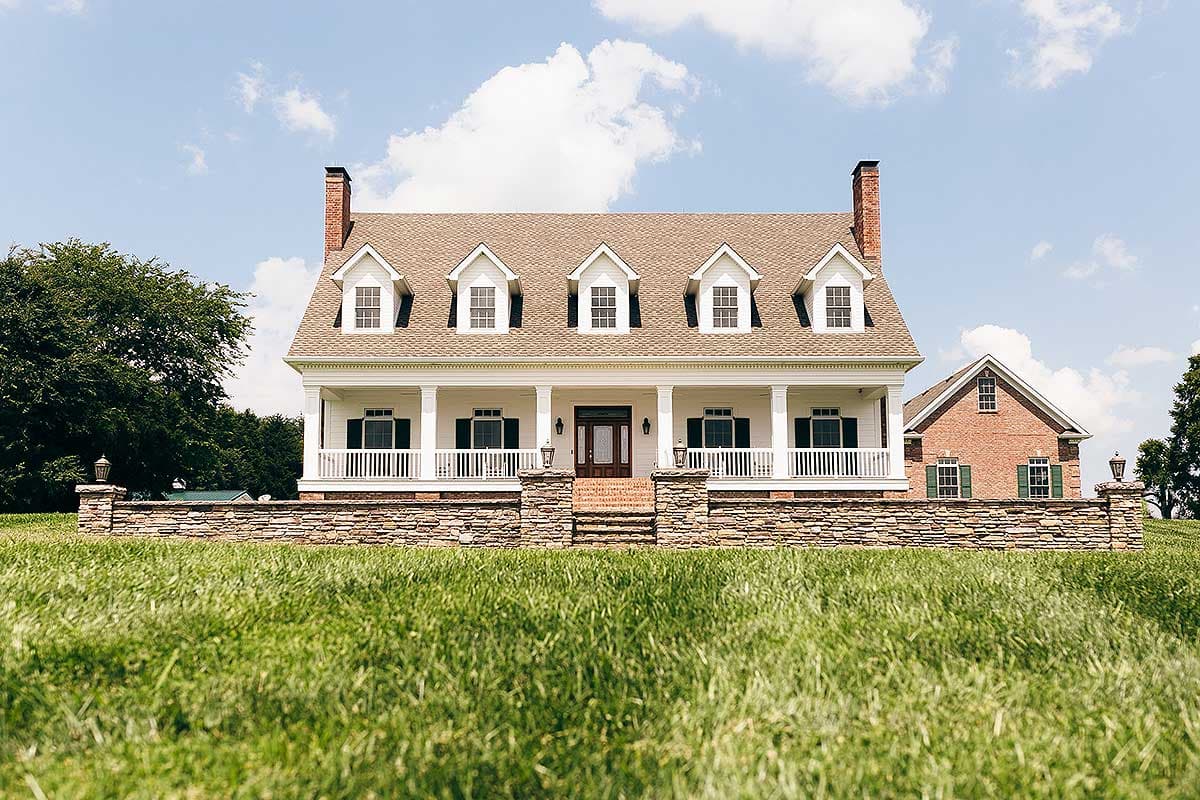 House plan exterior: Two-story home with full-width porch, five dormer windows, brick chimneys, and stone foundation wall.