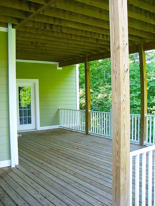 Wooden deck with white railings and support posts, attached to a house with green siding and a glass door.