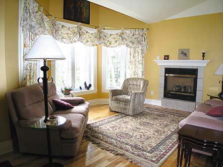 Living room interior with fireplace, bay window, and two seating areas with decorative curtains.