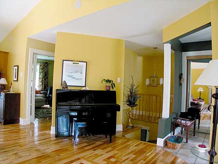 Interior view showing a black upright piano and a staircase with yellow railing and trim. A plant sits next to the piano.
