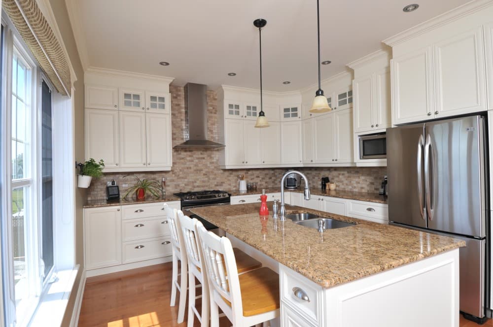 Kitchen with white cabinets, granite island, stainless steel appliances, tile backsplash, and large window.