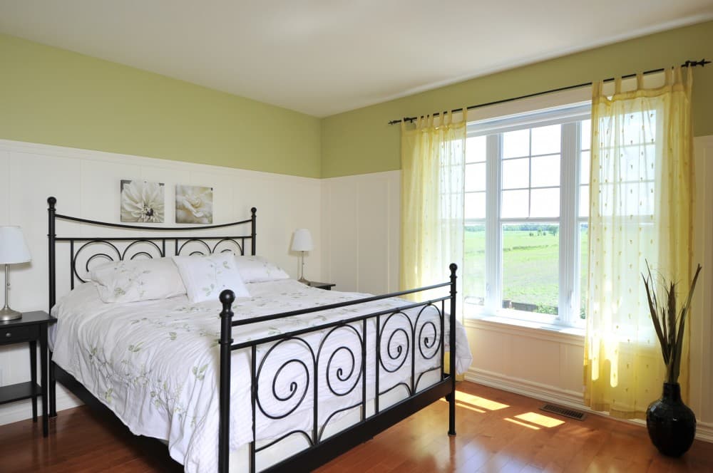 Bedroom with black metal bed frame, white bedding, wainscoting, and large window with yellow curtains overlooking a field.