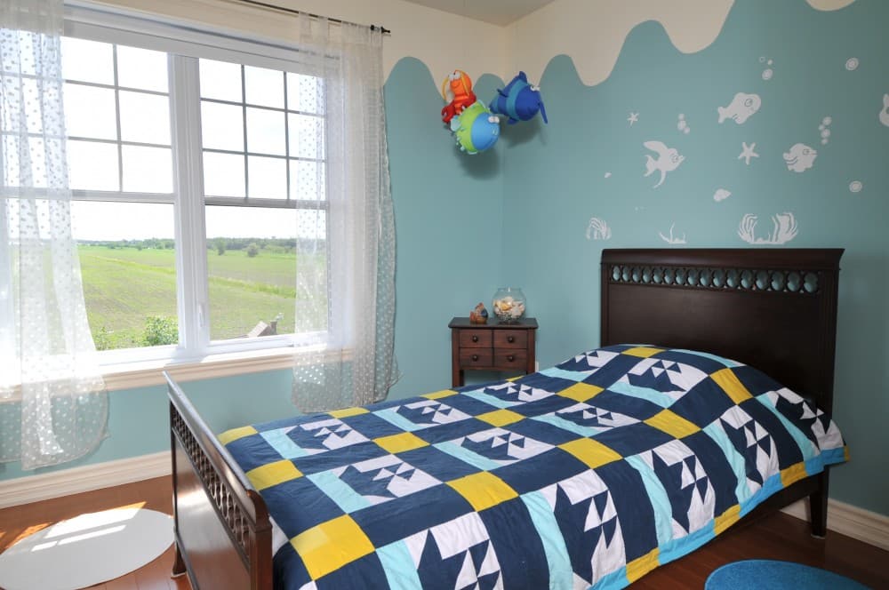 Children's bedroom with twin bed, blue quilt, and ocean-themed wall art. Large window overlooks a rural landscape.