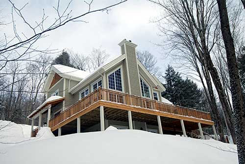 House plan exterior: Modern design with a large wraparound deck, prominent gabled dormers, and expansive A-frame windows.