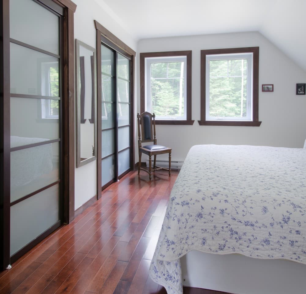 Bedroom with frosted glass sliding doors, wooden floors, and two windows overlooking trees. A chair sits near the windows.