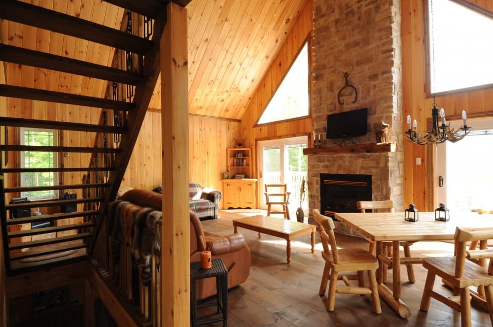 Log cabin interior with wood-paneled walls and ceiling, stone fireplace, dining set, and staircase.