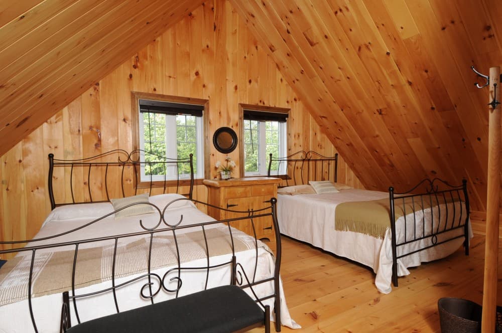 Interior view of a wood-paneled attic bedroom with two beds, a dresser, and two windows.