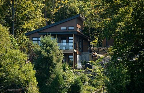 Modern house plan exterior with a steeply pitched roof, prominent windows, and a glass-railed deck nestled into a wooded hillside.