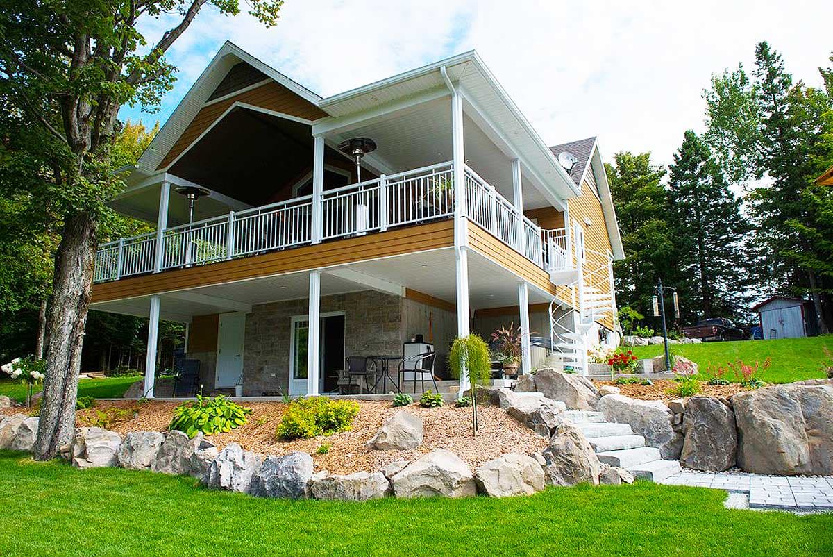 House plan exterior: Two-story home with a covered deck, stone foundation, and a spiral staircase.
