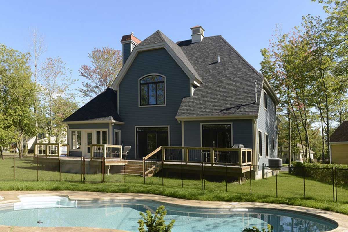 House plan exterior of a two-story home with a prominent gable, arched window, and a large wrap-around deck overlooking a pool.