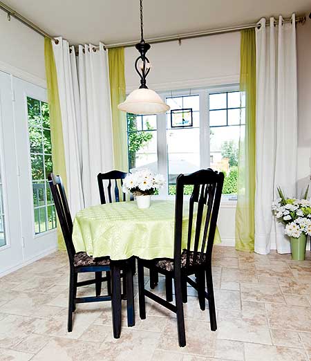 Dining area with round table, four chairs, pendant light, and large windows with white and green curtains.
