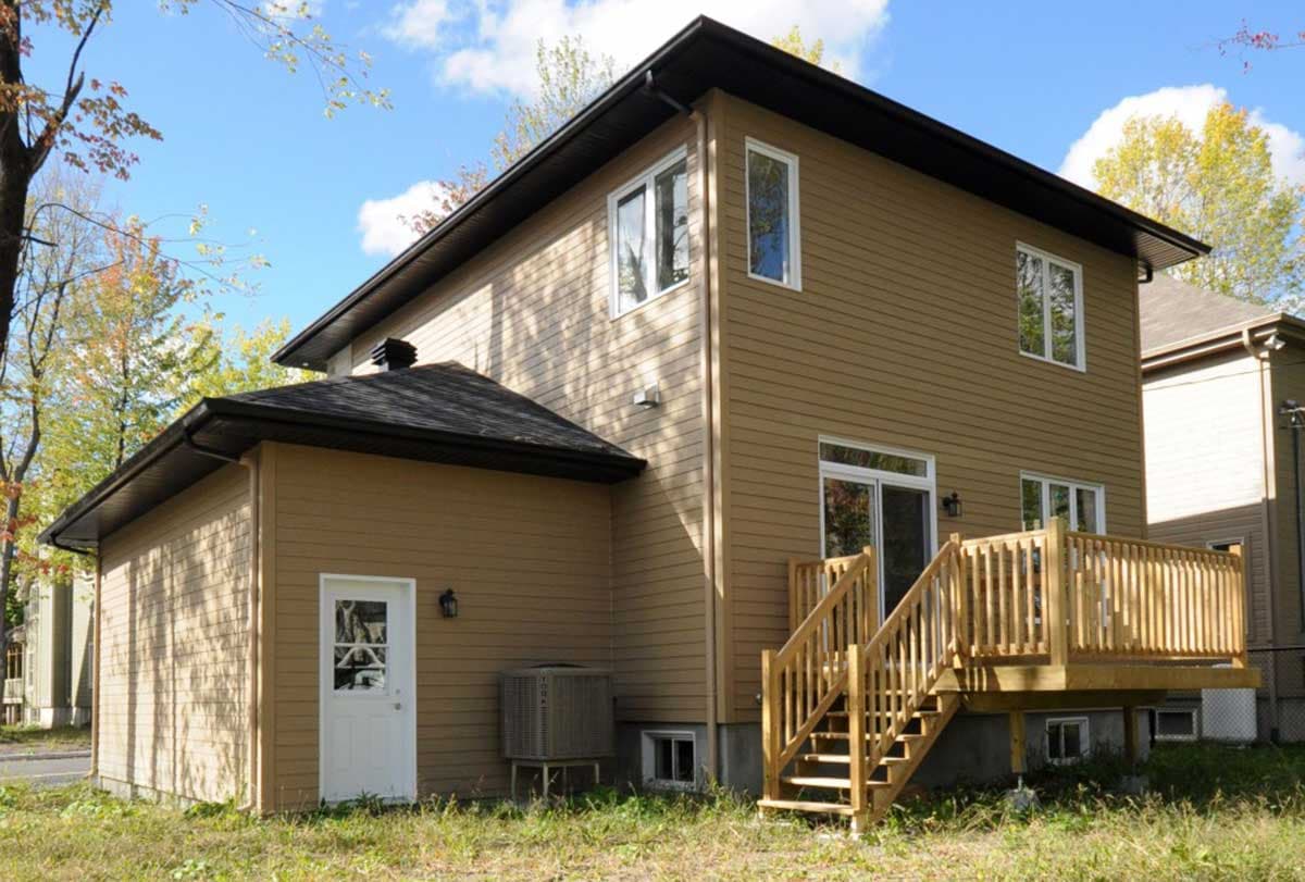 House plan exterior: Two-story home with tan siding, dark roof, a wooden deck with stairs, and a single front door.