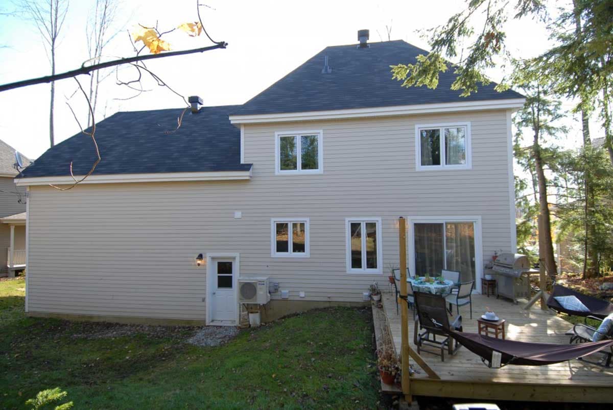 House plan exterior: two-story dwelling with beige siding, dark roof, multiple windows, a side door, and a rear deck with outdoor furniture.