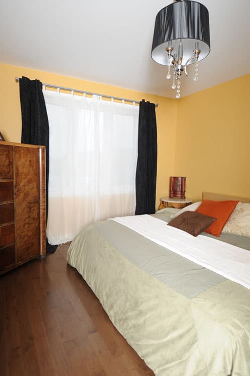 Bedroom with yellow walls, wood floor, large window with sheer and black curtains, wooden dresser, and chandelier.