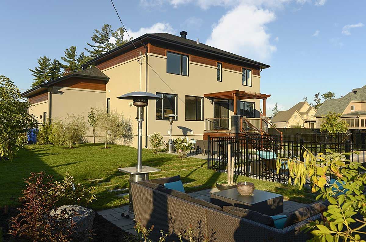 Two-story house plan exterior with stucco finish, wood trim accents, covered deck with pergola, and black metal fence.
