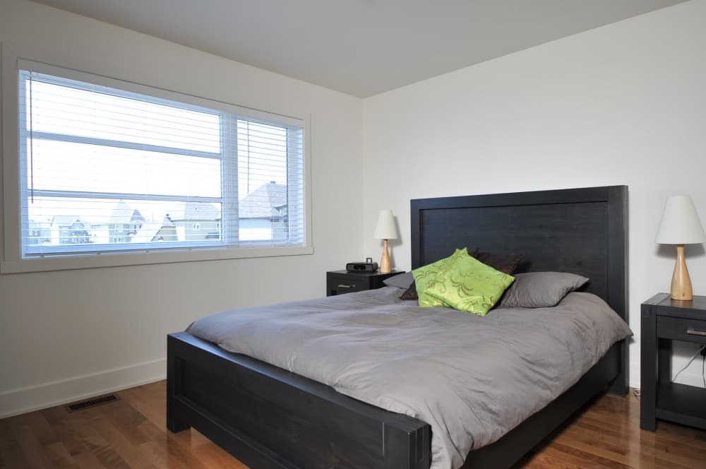 Bedroom with a king-size bed, dark wood headboard, and a large horizontal window with blinds overlooking houses.