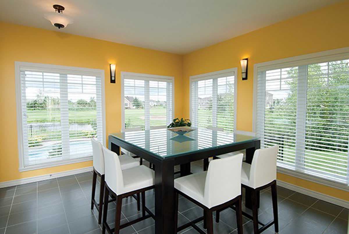 Dining area with a glass-top table, white bar stools, and large windows overlooking a backyard with a pool.