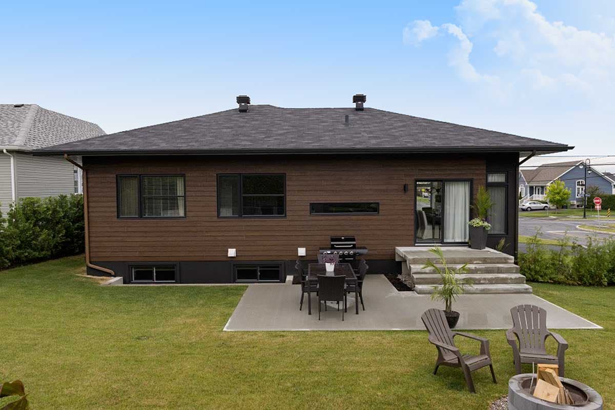 Exterior of a modern house plan featuring wood siding, a dark roof, a concrete patio with a dining set, and a concrete walkway with stairs.